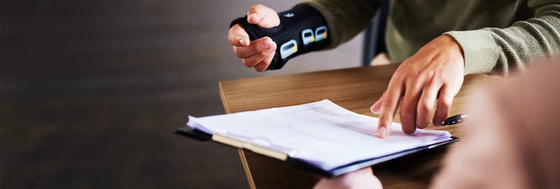 Two hands over a clipboard on a wooden table, one finger pointing at a document while a pen rests nearby; person wearing a green sweater.