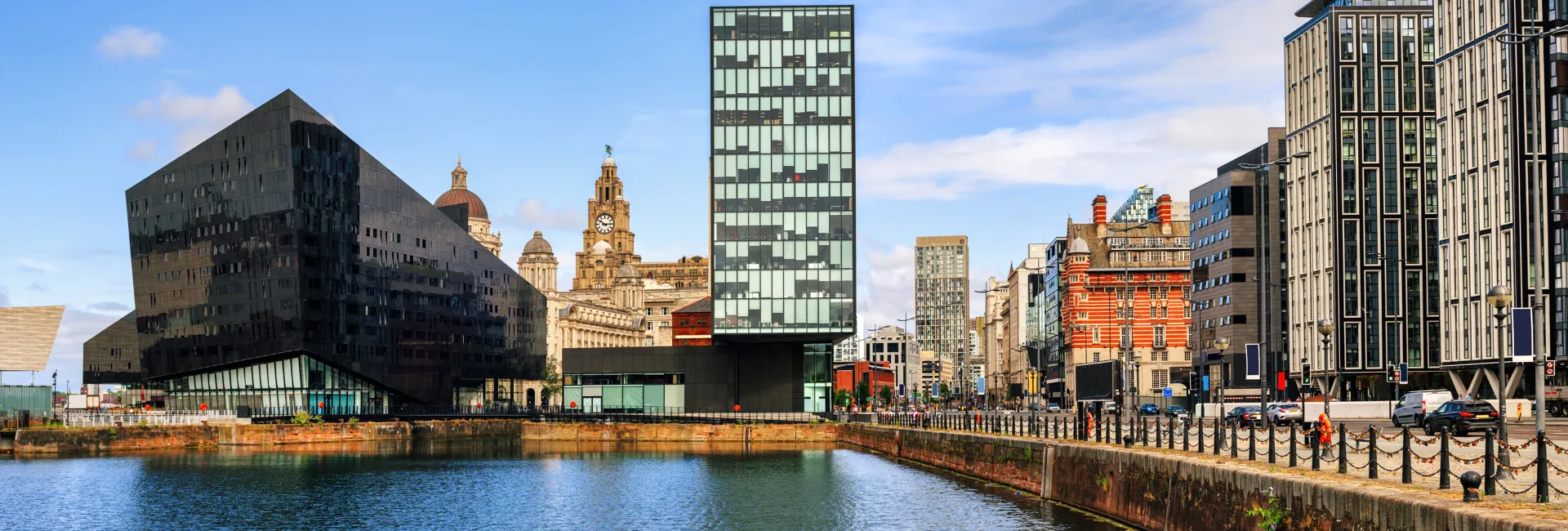 Panoramic city waterfront featuring sleek glass towers beside historic buildings, a calm river, and a pedestrian promenade under a blue sky.