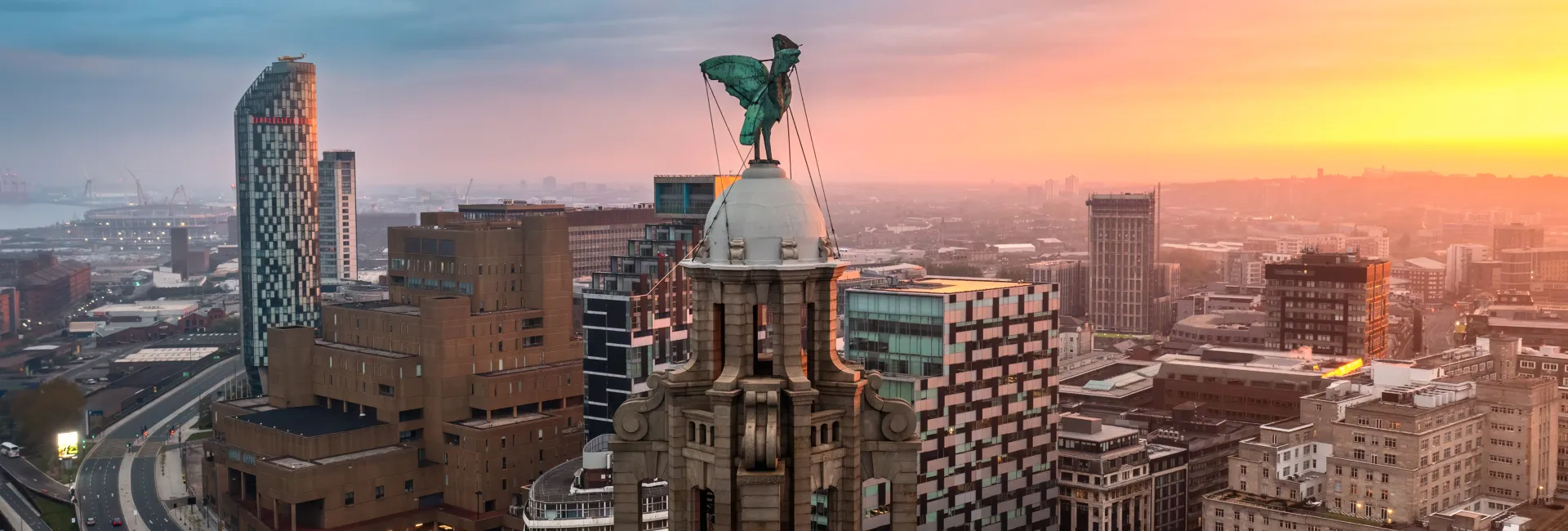 City skyline at sunset with a historic domed building topped by a green winged statue; surrounding modern glass towers glow orange in the haze.