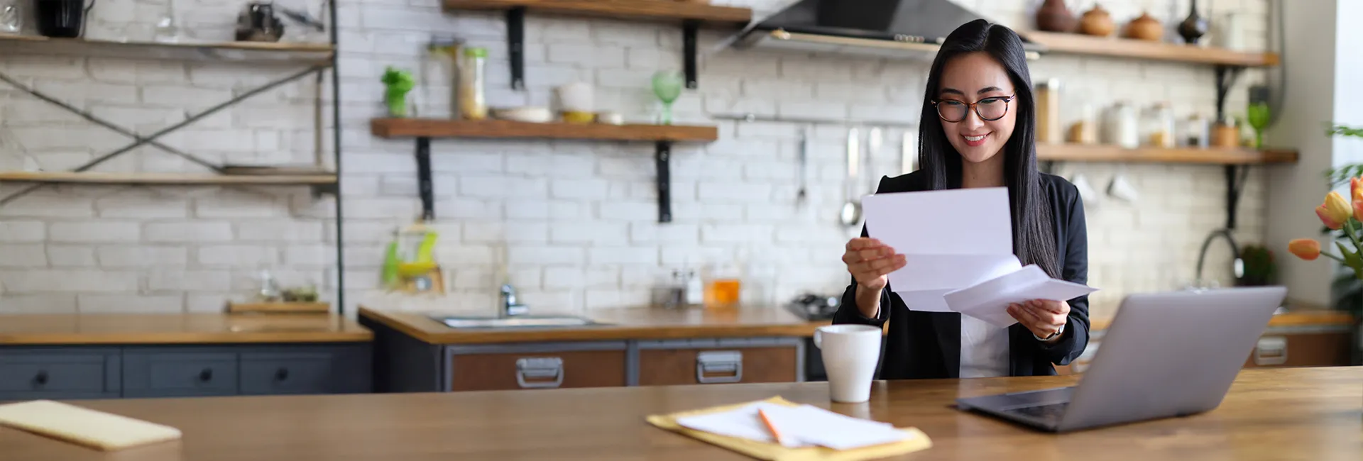 Person wearing glasses reads papers at a wooden kitchen island, with a laptop, mug, and tulips nearby; shelves and a brick wall in the background.