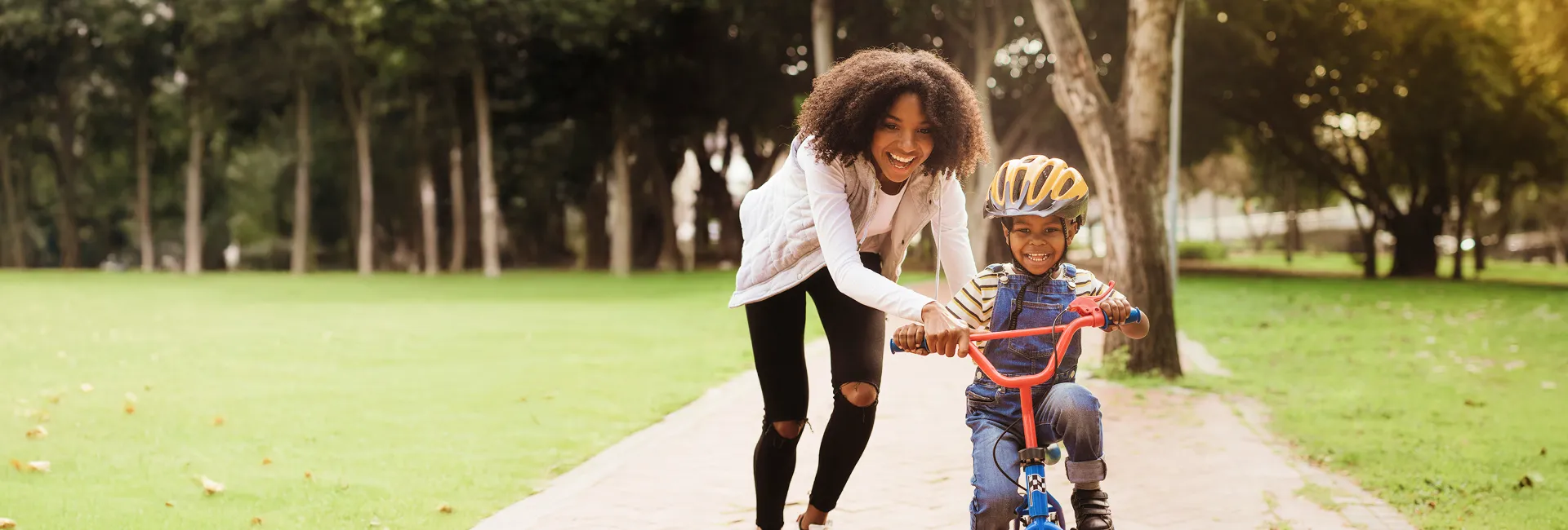 A smiling adult with curly hair helps a child wearing a helmet ride a small red bicycle along a sunlit park path lined with trees.