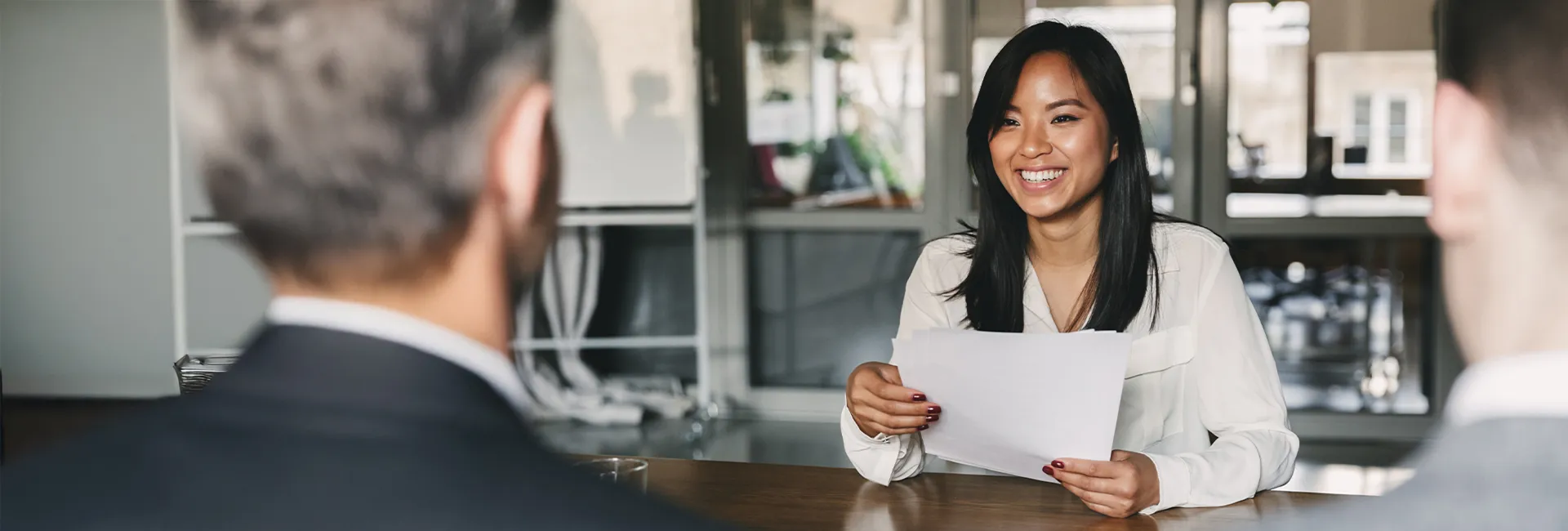 Smiling person with long dark hair in a white blouse sits at a table, holding papers while two blurred colleagues listen in an office.
