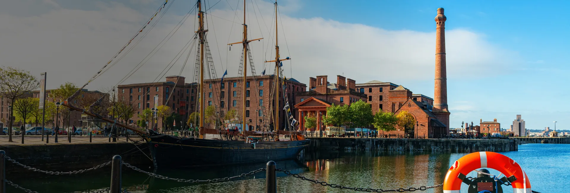 Harbor with tall-masted ship, red-brick warehouses, a brick chimney, trees along the quay, reflections in water, and an orange lifebuoy in the foreground.