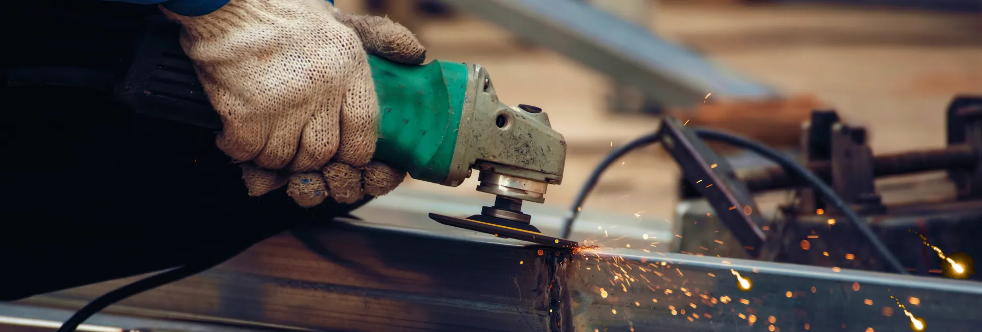 Close-up of gloved hands guiding a green angle grinder as sparks fly while cutting a metal beam.