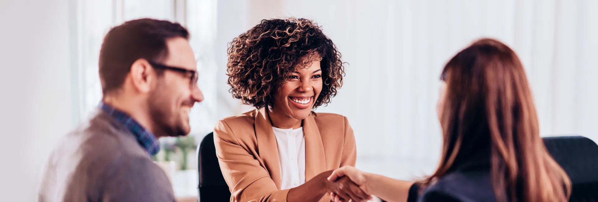 Three colleagues in a bright office; the center person with curly hair smiles while shaking hands with the person on the right, as a third person watches.