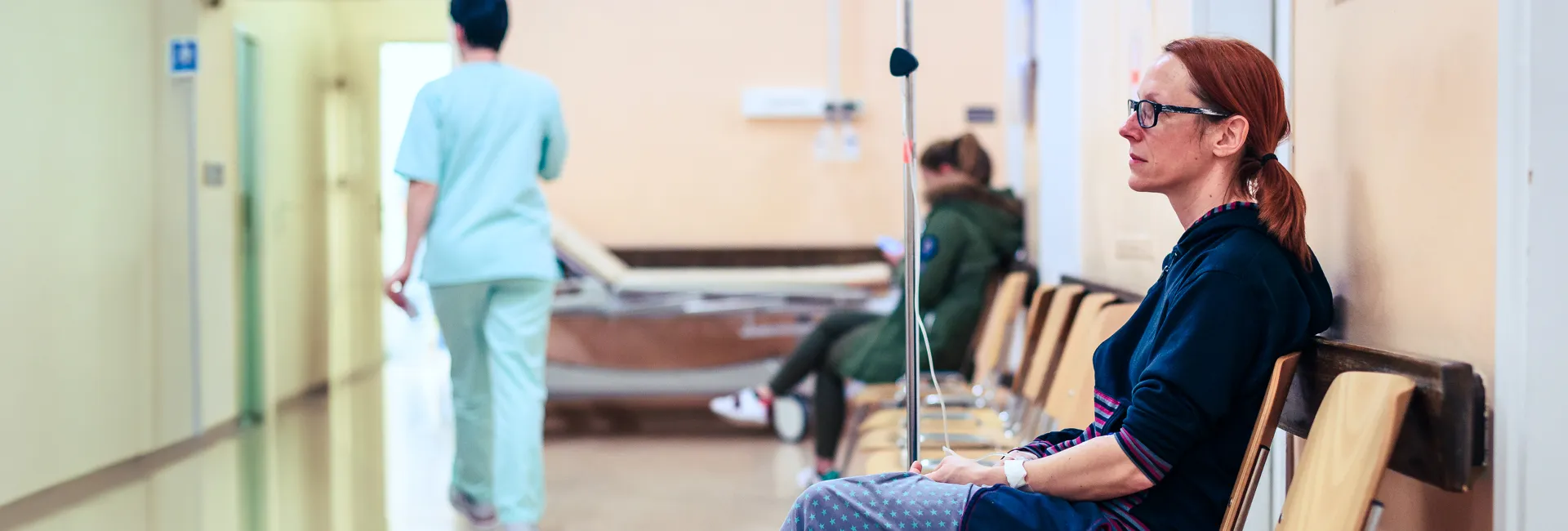 Hospital corridor: a nurse pushes a stretcher while a patient waits in the background; a red-haired person with glasses sits on a bench to the right.