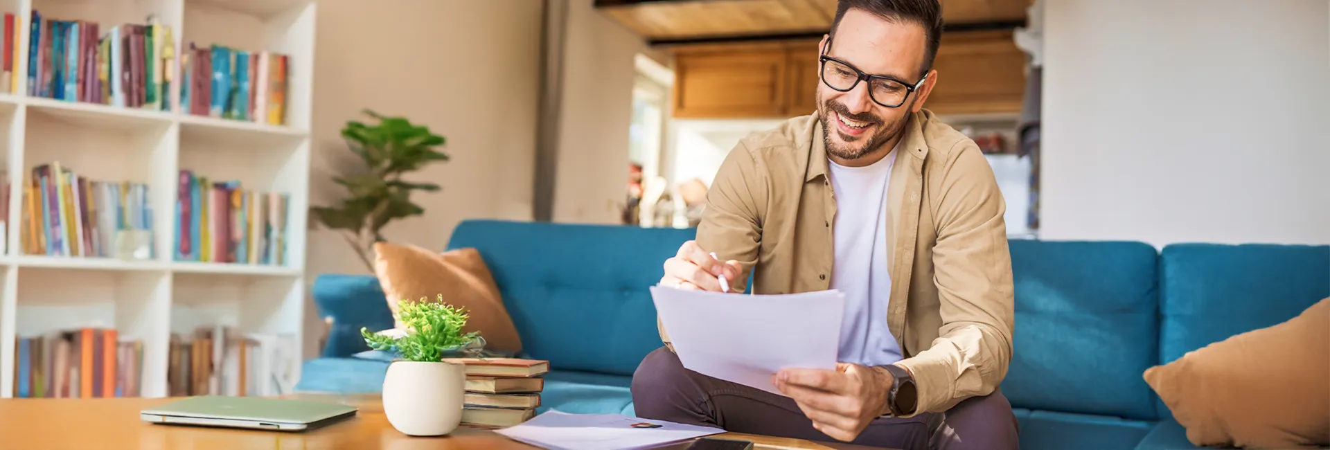 Person with short dark hair and glasses sits on a blue sofa, smiling while reviewing papers; a coffee table with a laptop, plant, and books nearby.