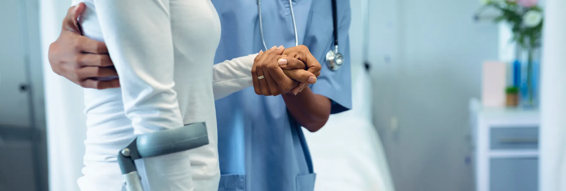 A healthcare professional in blue scrubs holds the hand of a patient in white, offering support in a hospital corridor; a forearm crutch rests nearby.