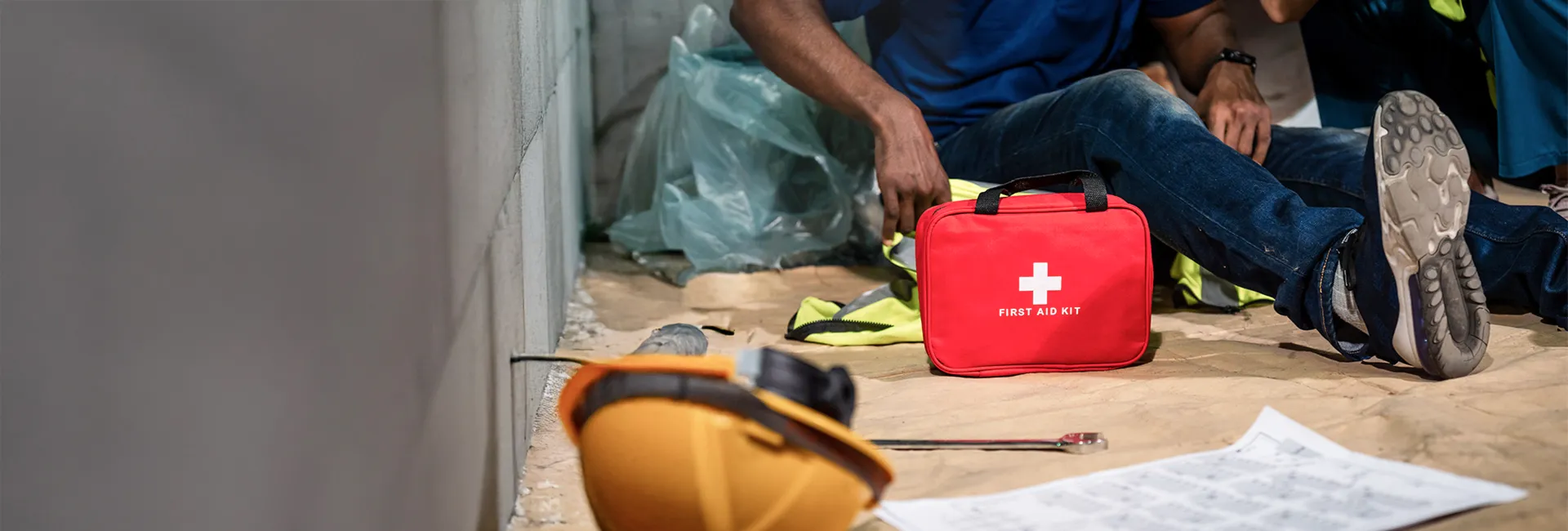 Red first aid kit with a white cross on the floor beside a person wearing jeans; scattered safety gear and a hard hat nearby.
