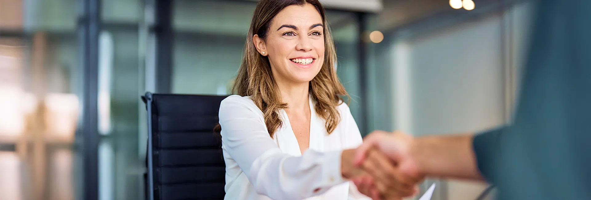 Smiling person with shoulder-length light brown hair in a white blouse shakes hands with another person in a modern office as glass walls blur in the background.