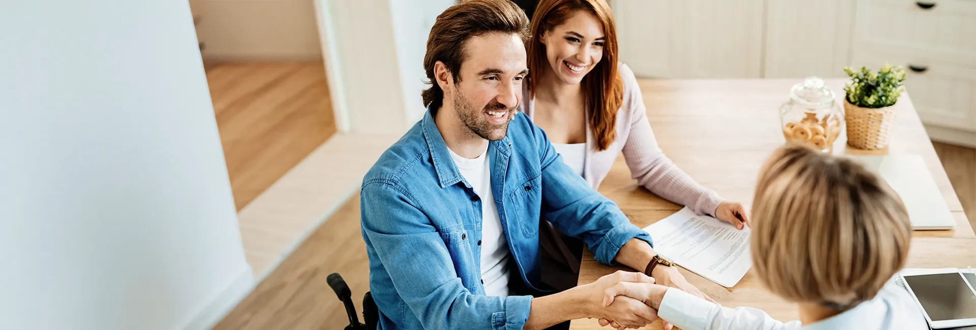 Smiling couple in a bright kitchen shake hands with a professional across a wooden table; documents, a cookie jar, and a plant nearby.