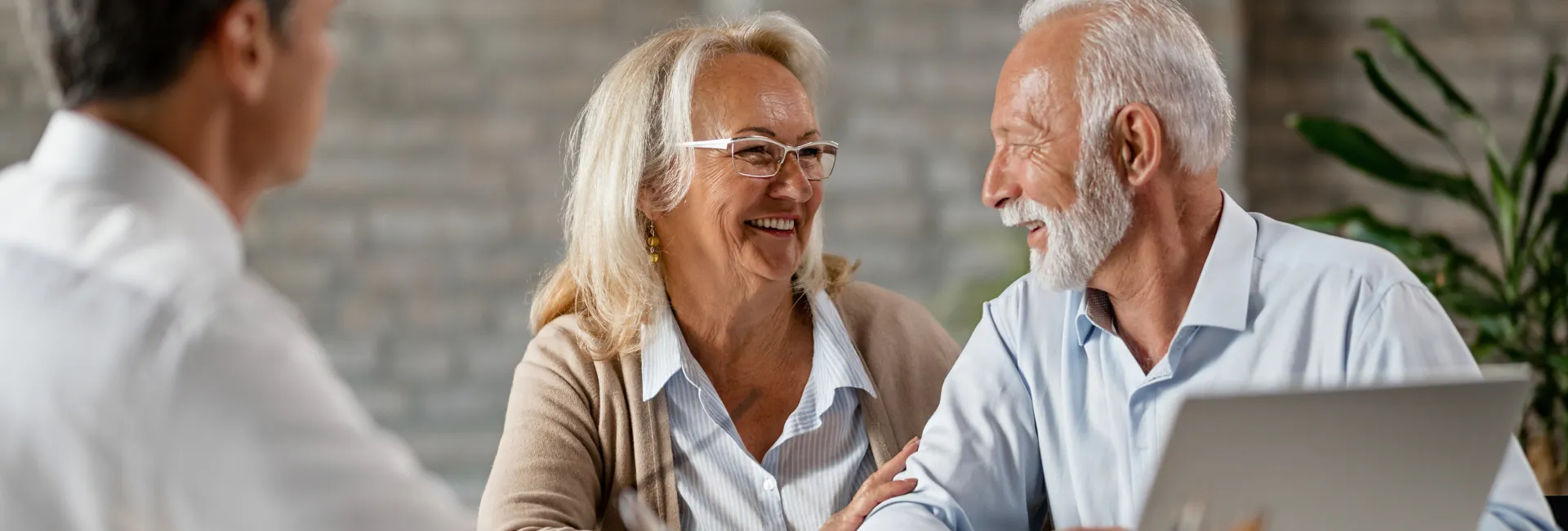 Two seniors in a bright office chat around a laptop; smiling at each other while a blurred colleague sits at left, with plants and brick wall in background.