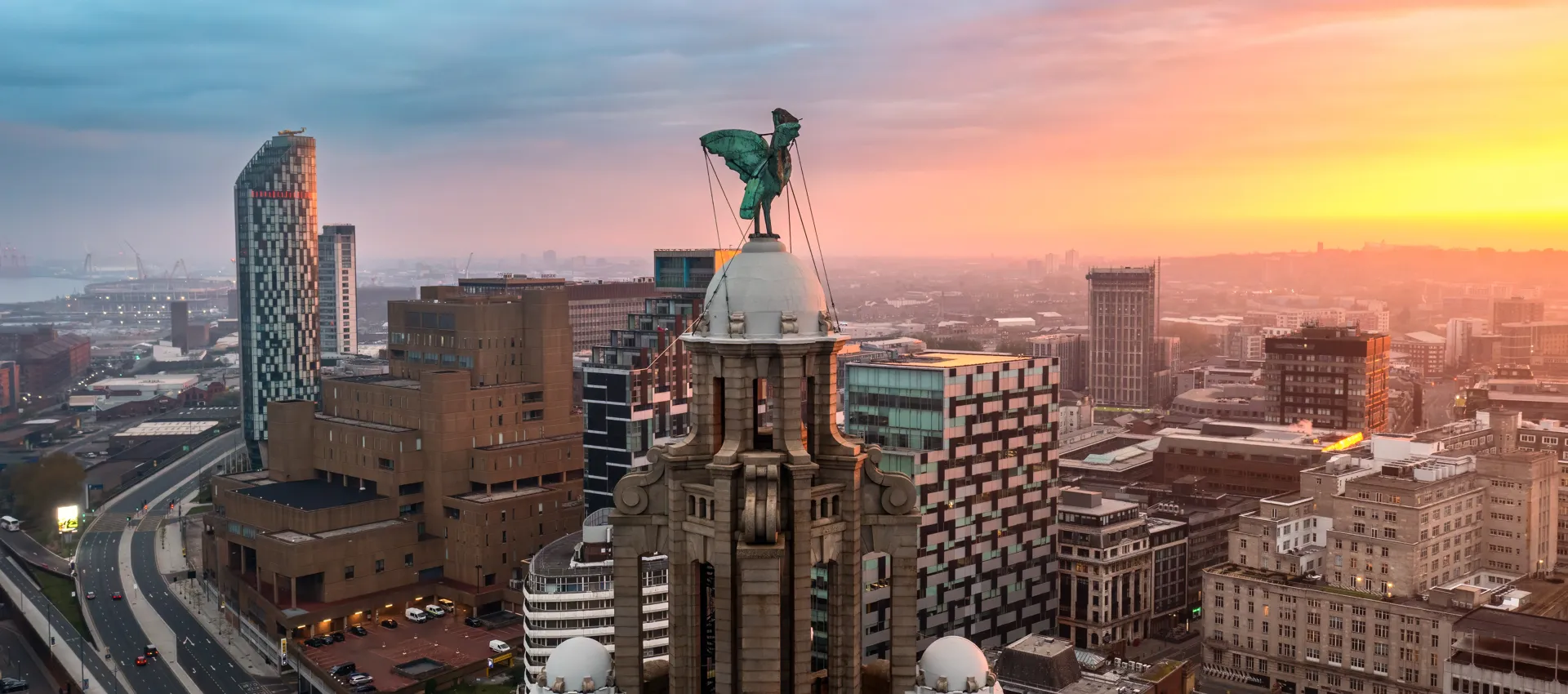 An aerial cityscape at sunset, with a central ornate dome crowned by a green winged statue, surrounded by modern towers and warm orange light.