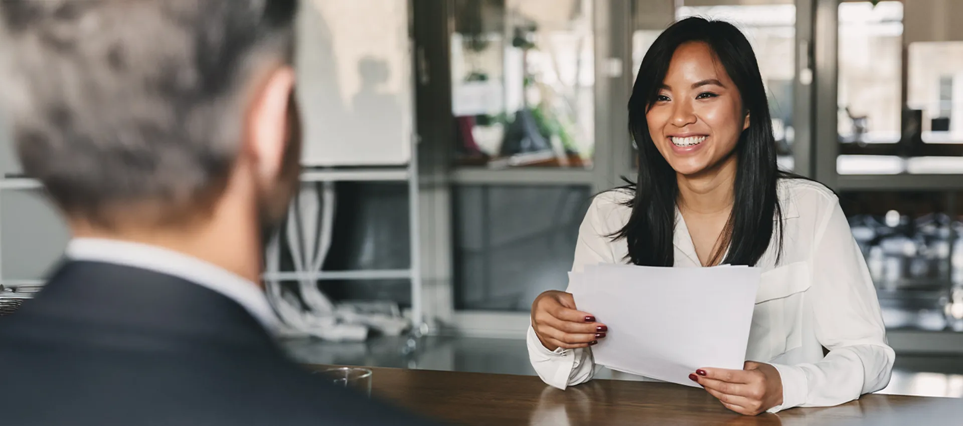 Smiling person with long dark hair in a white blouse sits at a table, holding papers while two blurred colleagues listen in an office.