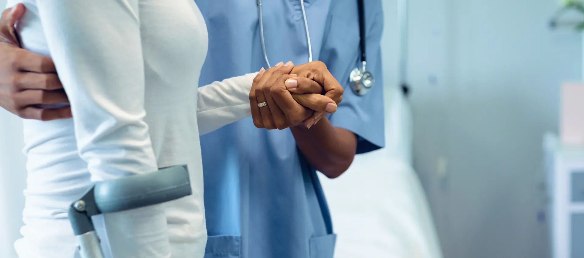 A healthcare professional in blue scrubs holds the hand of a patient in white, offering support in a hospital corridor; a forearm crutch rests nearby.
