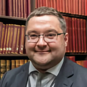 Close-up portrait of a person wearing glasses and a suit, smiling, with shelves of red and brown books in the background. profile photo