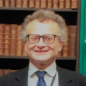 Smiling person with curly gray hair and black-rimmed glasses, wearing a suit and blue tie, in front of a bookshelf of brown leather-bound books. profile photo