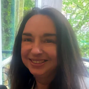 Close-up of a smiling person with long dark hair, facing the camera; a bright window with green plants visible in the background. profile photo
