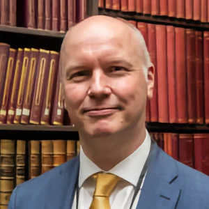 A person with a shaved head wearing a blue suit and yellow tie stands in front of a bookshelf filled with red and brown leather-bound volumes, smiling. profile photo