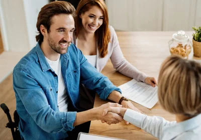 Person in a wheelchair shakes hands with another across a wooden table; a smiling colleague sits beside them, with documents, a jar of cookies and a plant nearby.