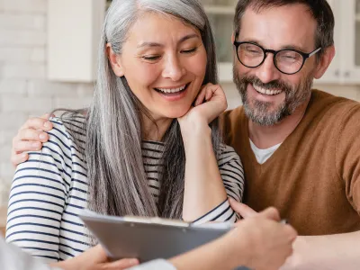 Two adults smile while looking at a clipboard in a kitchen. One has long gray hair and a striped shirt; the other wears glasses and a brown sweater.