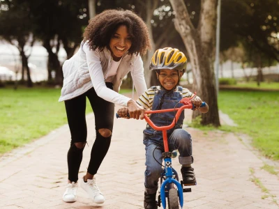 An adult smiles while helping a child ride a blue bike with red handlebars along a park path; child wears a yellow helmet, striped shirt, overalls.