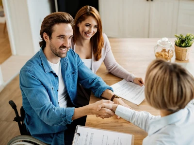 A person in a denim shirt sits in a wheelchair and shakes hands with a professional across a wooden table, while another person smiles beside papers on table.