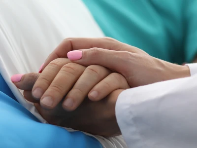Two hands gently clasped in a comforting hold, one with pink nail polish, in a medical setting.