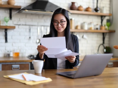 A person with long dark hair and glasses sits at a wooden kitchen counter, reading papers with a laptop nearby and a white mug on the table.