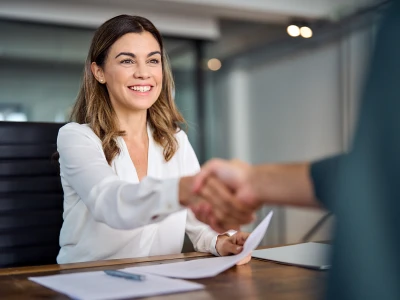 Smiling person in a white blouse shakes hands across a desk in a modern office, with papers on the table.
