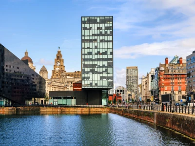Cityscape with a tall glass tower at center, domed clock tower in the background, red brick buildings to the right, and a water canal in foreground.