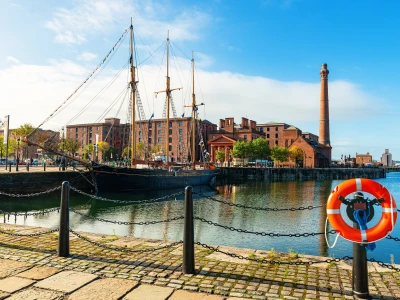 Historic sailing ship moored at a brick warehouse quay, with a tall chimney, calm water, and a bright orange lifebuoy on the railing in the foreground.