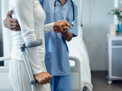 A healthcare professional in blue scrubs consoles a patient using a forearm crutch, holding their hand; stethoscope visible.