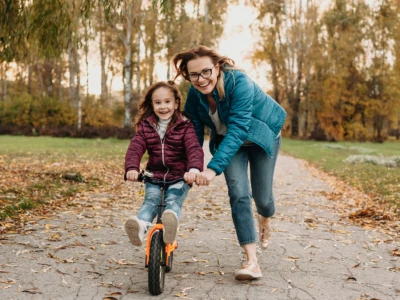 An adult helps a child ride a small orange bike along a leaf-strewn park path, with autumn trees in the background.