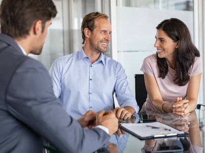 Three colleagues in business attire sit at a glass conference table in a bright office, smiling during a meeting.