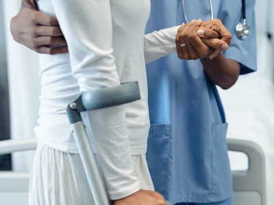A clinician in blue scrubs holds a patient's hand while the patient uses forearm crutches in a hospital setting.