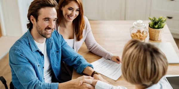 Smiling couple sits at a wooden table with a document, shaking hands with a third person; a cookie jar and plant on the table in a bright kitchen.