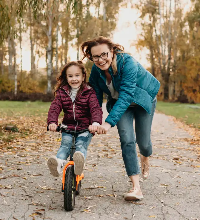 An adult helps a child ride a small orange bike along a leaf-strewn park path in autumn; both are smiling.