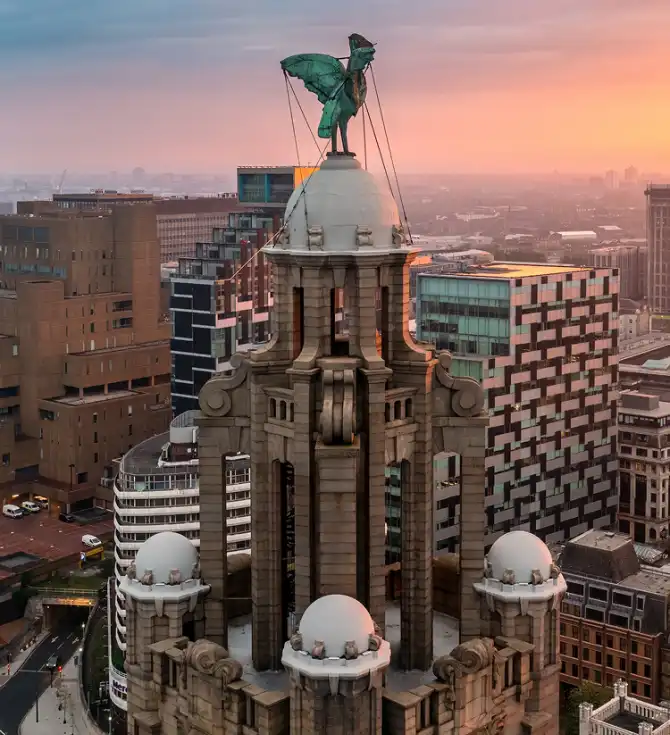 Historic tower with a winged statue atop a dome, framed by surrounding modern buildings as a warm pink-orange sunset lights the city.