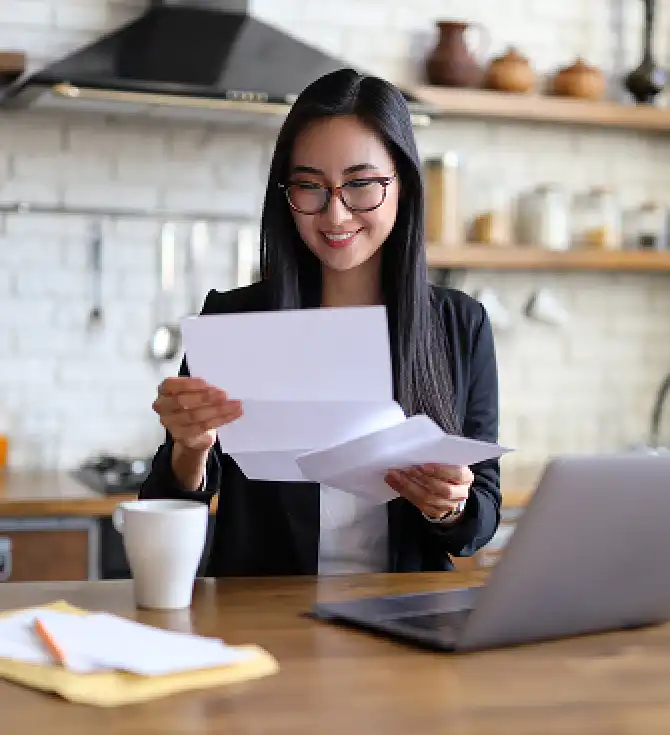 Person with long dark hair and glasses smiles while reading papers at a wooden table beside an open laptop in a bright kitchen.