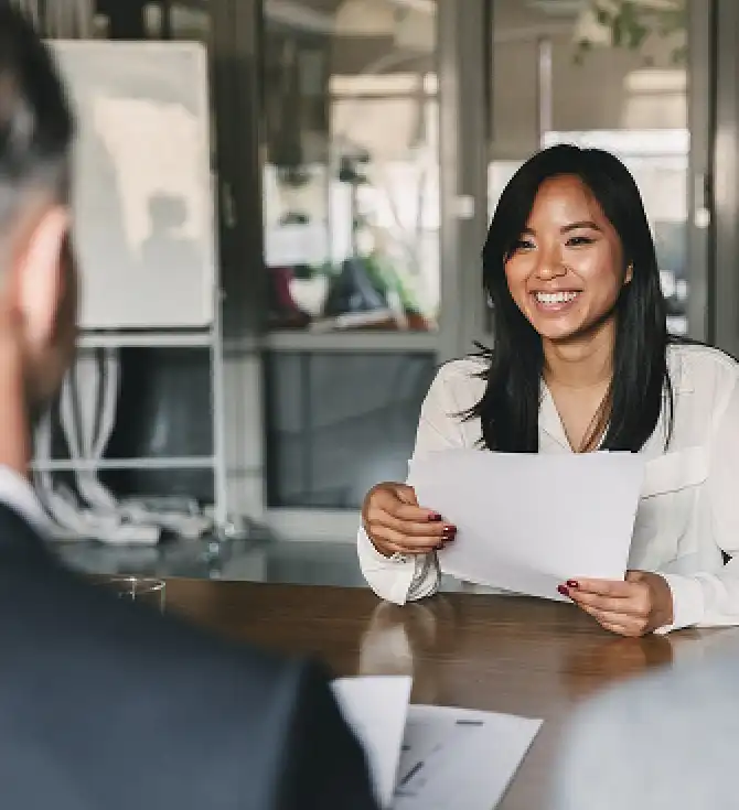 Smiling person with long dark hair in a white blouse sits at a wooden table, holding a sheet of paper across from another person in a modern office.