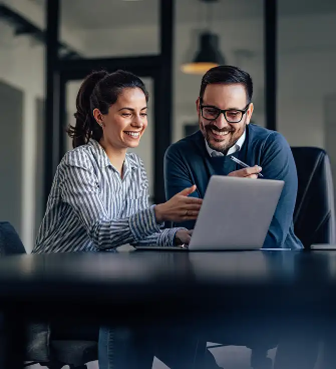 Two colleagues collaborate at a desk with a laptop in a modern office, smiling as they discuss a project.