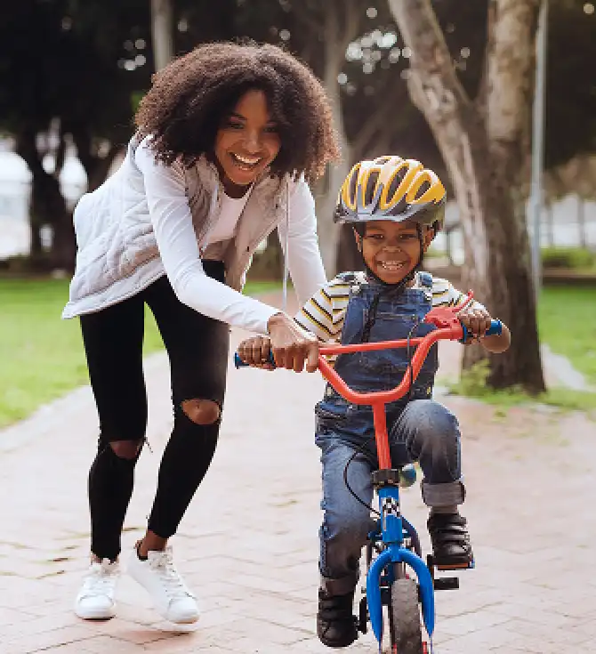 A smiling adult guides a child wearing a yellow helmet as they ride a blue bike with red handlebars along a park path.