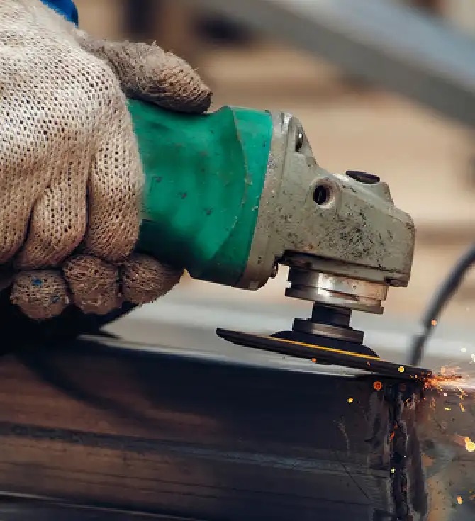 Close-up of a gloved hand using a green angle grinder to grind metal on a work surface, sparks flying around the cutting area.