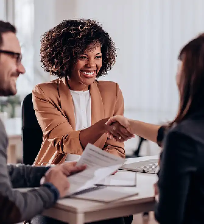 Three people sit at a bright office desk; a person with curly hair and a tan blazer smiles while shaking hands with another person, with papers and a laptop on the table.