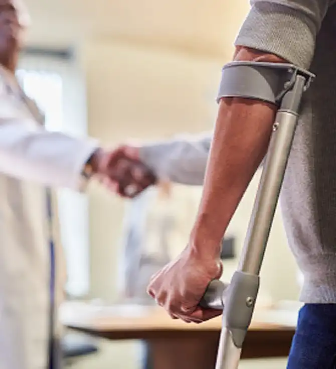 Person using a forearm crutch in the foreground; two colleagues shake hands in a clinical setting with a softly blurred background.