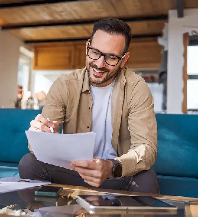 A person with short dark hair and glasses sits on a blue couch, smiling while reviewing papers with a pen in hand in a warm, modern living space.
