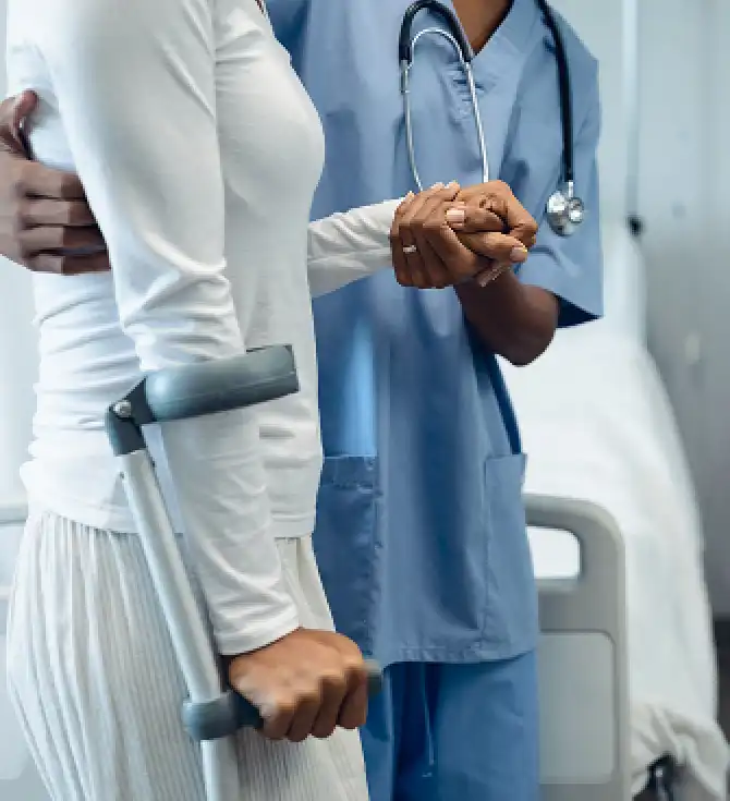 A healthcare professional in blue scrubs holds the hand of a patient using a forearm crutch in a hospital corridor.