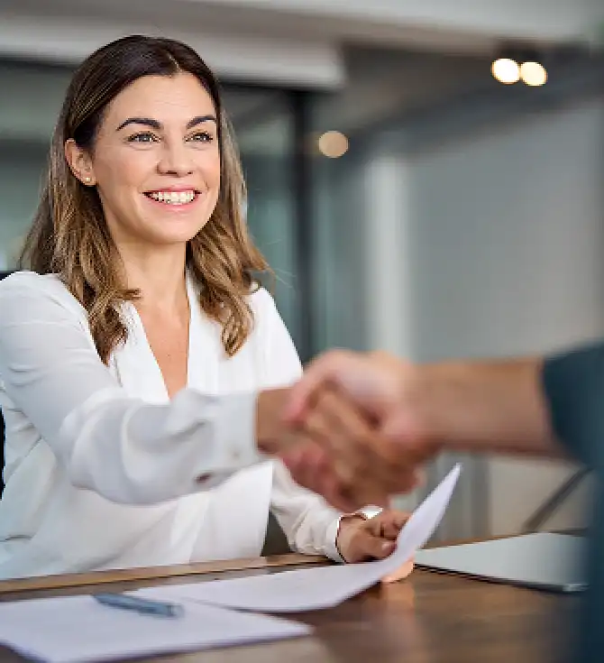 Person with shoulder-length hair and white blouse smiles while shaking hands across a desk in a modern office; papers and a laptop on the desk.