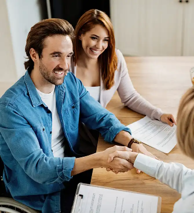 Two people shake hands across a table with documents; one in a blue denim shirt, the other in a light cardigan, smiling, with a third person partly visible.
